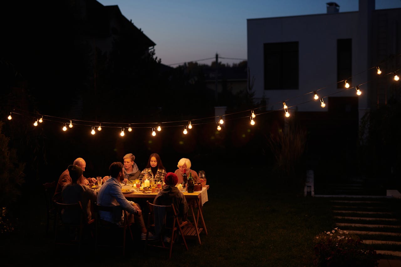 A family enjoys an evening meal outdoors under decorative string lights in a garden setting.