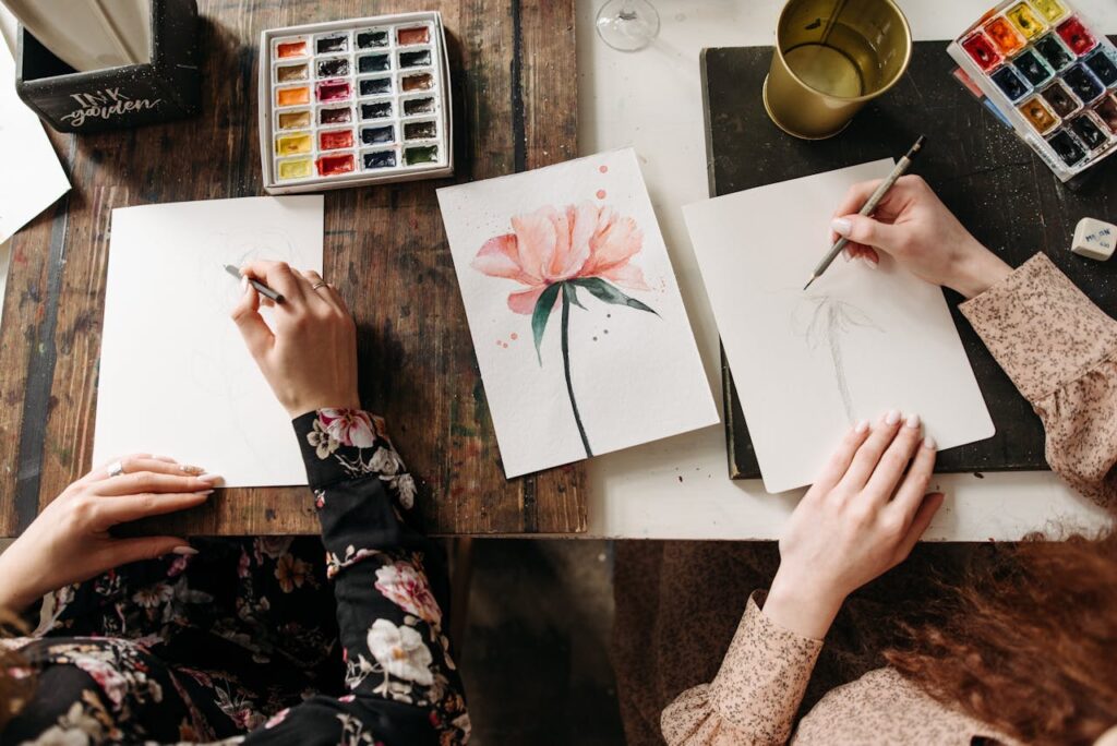 Overhead view of artists drawing floral designs with watercolors and pencils, indoors.